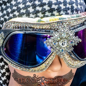 Puede incluir: Gafas protectoras con lentes reflectantes azul oscuro, montura negra y un broche central plateado con cristales transparentes. Los bordes est&aacute;n decorados con filas de peque&ntilde;os diamantes de imitaci&oacute;n y una correa el&aacute;stica de color dorado.