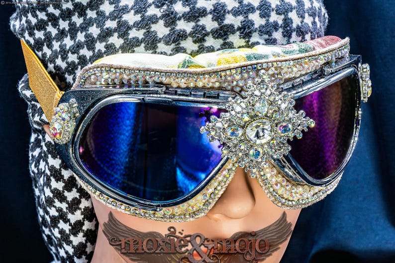 Puede incluir: Gafas protectoras con lentes reflectantes azul oscuro, montura negra y un broche central plateado con cristales transparentes. Los bordes est&aacute;n decorados con filas de peque&ntilde;os diamantes de imitaci&oacute;n y una correa el&aacute;stica de color dorado.