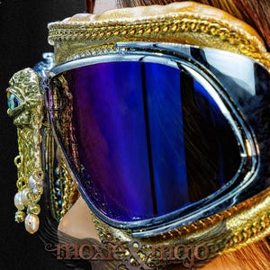 May include: Goggles featuring reflective blue lenses and a gold-toned frame with intricate braided trim. A gold-colored eye-shaped ornament is attached to the side, adorned with dangling white and pink pearls. A brown strap secures the eyewear.