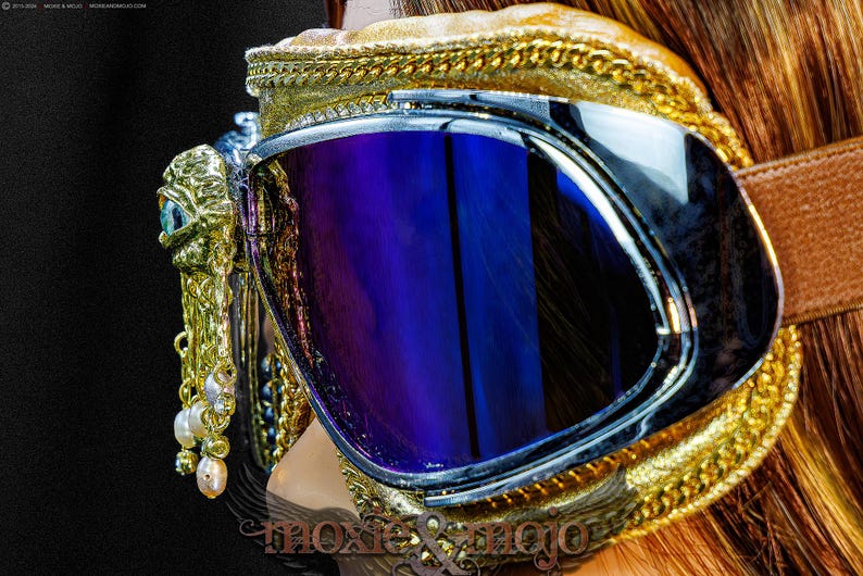 May include: Goggles featuring reflective blue lenses and a gold-toned frame with intricate braided trim. A gold-colored eye-shaped ornament is attached to the side, adorned with dangling white and pink pearls. A brown strap secures the eyewear.