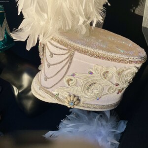 May include: White drum major hat with white feathers, white and silver embellishments, and a white and silver design. The hat is on a black surface.