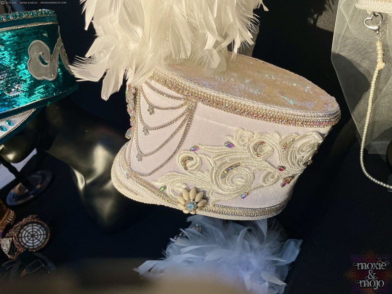 May include: White drum major hat with white feathers, white and silver embellishments, and a white and silver design. The hat is on a black surface.