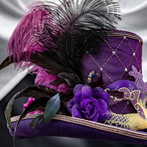 May include: A purple top hat featuring a diamond-patterned mesh overlay with small blue accents. The hat is adorned with a large purple fabric rose, black and purple feathers, and a decorative purple and gold masquerade mask on the side.