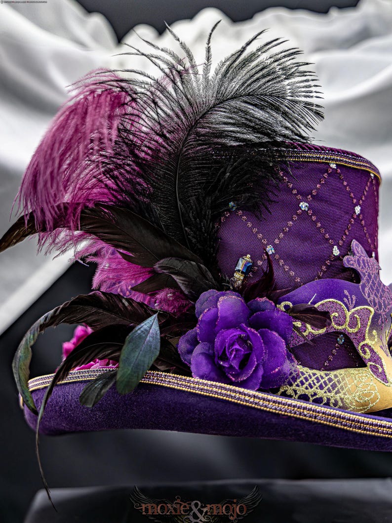 May include: A purple top hat featuring a diamond-patterned mesh overlay with small blue accents. The hat is adorned with a large purple fabric rose, black and purple feathers, and a decorative purple and gold masquerade mask on the side.