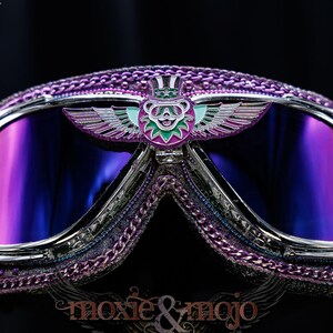Puede incluir: Gafas protectoras de color morado y cromo con un emblema central de un oso alado con sombrero de copa. Las lentes tienen un tono reflectante morado y azul. El marco est&aacute; decorado con adornos morados y peque&ntilde;os detalles blancos en los bordes.