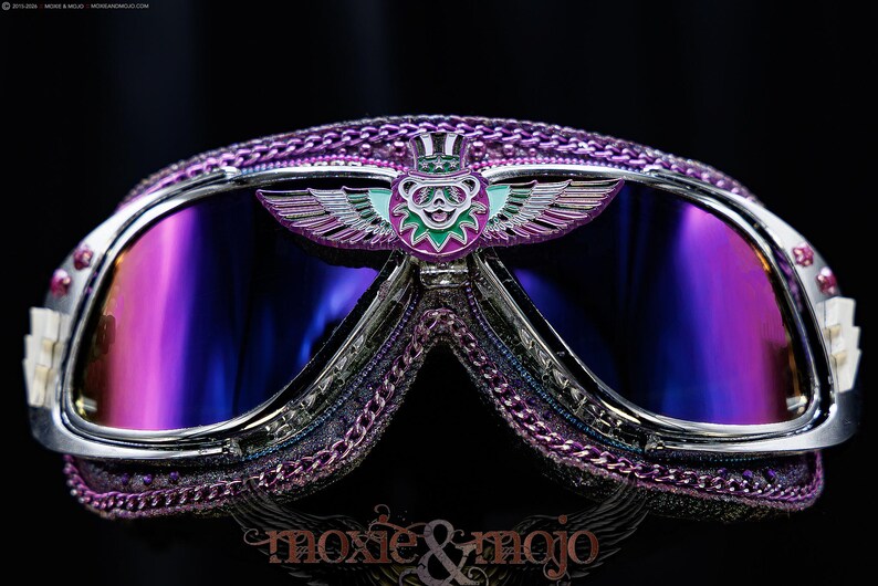 Puede incluir: Gafas protectoras de color morado y cromo con un emblema central de un oso alado con sombrero de copa. Las lentes tienen un tono reflectante morado y azul. El marco est&aacute; decorado con adornos morados y peque&ntilde;os detalles blancos en los bordes.