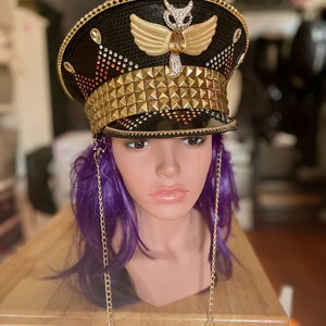 May include: A black and gold embellished hat with a gold owl design and a row of gold pyramid-shaped studs. The hat is on a mannequin with purple hair and a gold chain necklace.