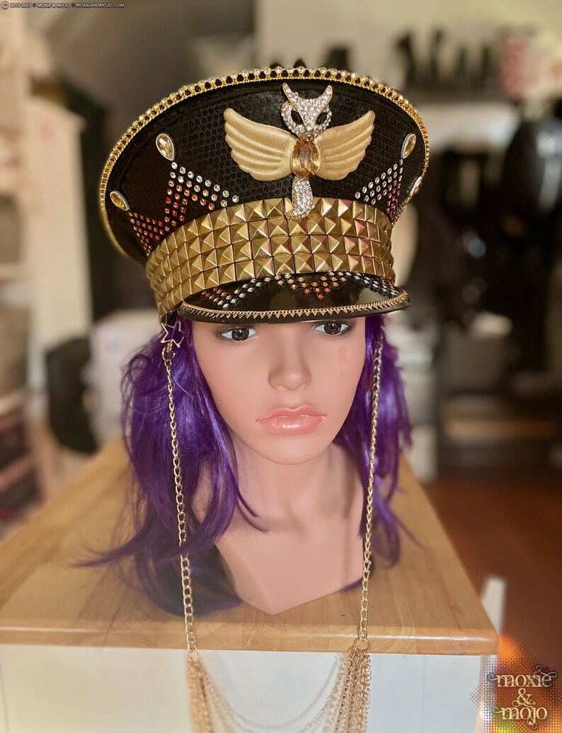 May include: A black and gold embellished hat with a gold owl design and a row of gold pyramid-shaped studs. The hat is on a mannequin with purple hair and a gold chain necklace.