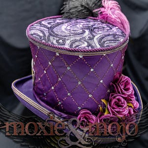 May include: A purple top hat featuring a diamond-patterned lattice overlay with embedded clear crystals. The crown is adorned with a swirling patterned fabric and black and purple feathers. A cluster of purple fabric roses sits on the brim.