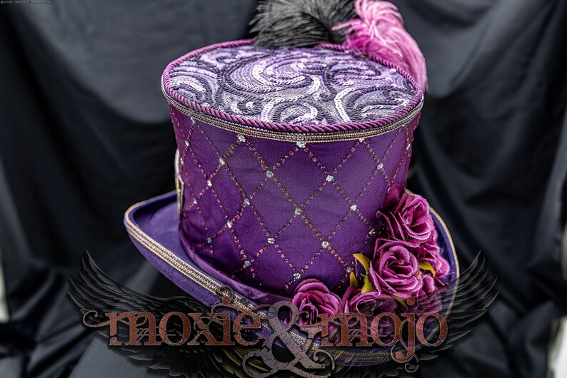 May include: A purple top hat featuring a diamond-patterned lattice overlay with embedded clear crystals. The crown is adorned with a swirling patterned fabric and black and purple feathers. A cluster of purple fabric roses sits on the brim.