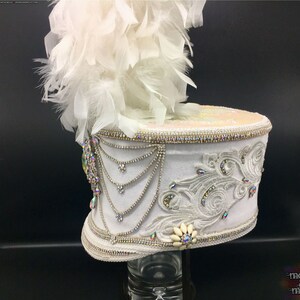 May include: White drum major hat with a white feather plume, embellished with rhinestones and a white flower. The hat is on a black background.