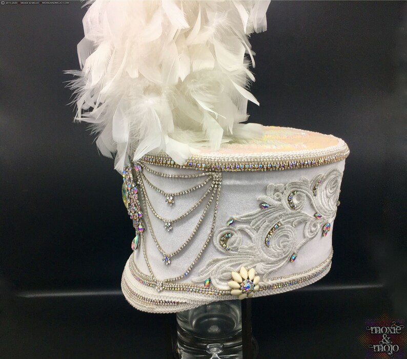 May include: White drum major hat with a white feather plume, embellished with rhinestones and a white flower. The hat is on a black background.