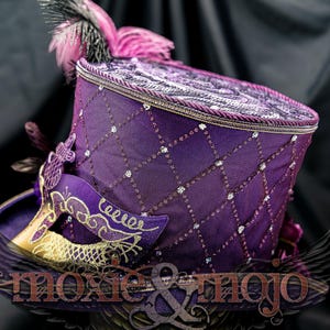 May include: A purple top hat featuring a diamond-patterned overlay with embedded clear crystals, a braided trim along the top edge, and a purple and gold masquerade mask attached to the side. Pink and black feathers accent the top of the hat.