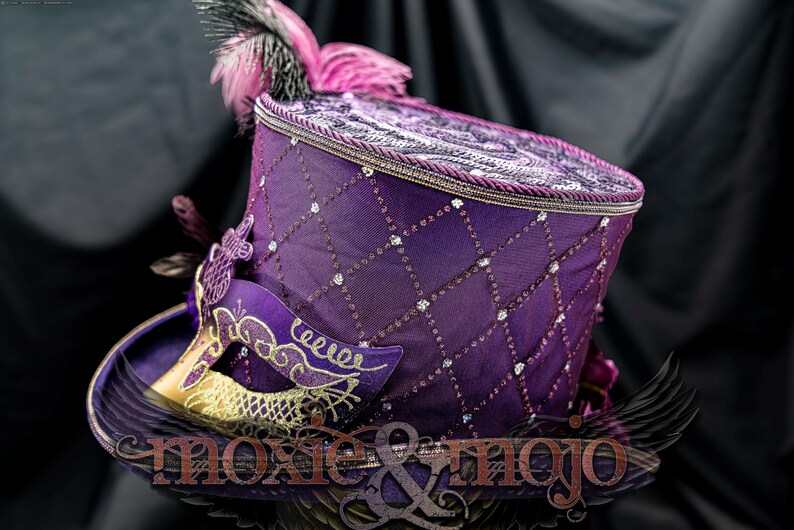 May include: A purple top hat featuring a diamond-patterned overlay with embedded clear crystals, a braided trim along the top edge, and a purple and gold masquerade mask attached to the side. Pink and black feathers accent the top of the hat.