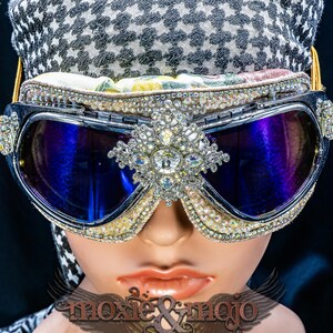 Puede incluir: Gafas protectoras con lentes azul oscuro y una montura densamente incrustada con brillantes transparentes. Un gran adorno de pedrer&iacute;a est&aacute; centrado entre las lentes. Las gafas se llevan sobre una tela con estampado de pata de gallo en blanco y negro.