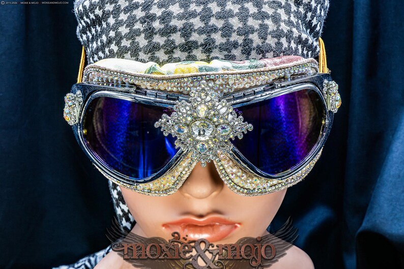 Puede incluir: Gafas protectoras con lentes azul oscuro y una montura densamente incrustada con brillantes transparentes. Un gran adorno de pedrer&iacute;a est&aacute; centrado entre las lentes. Las gafas se llevan sobre una tela con estampado de pata de gallo en blanco y negro.