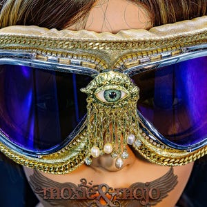 May include: Goggles with dark purple lenses and a silver frame, featuring a gold-toned trim with chain accents. A central gold ornament depicts an eye with dangling pearl and crystal charms. The text moxie & mojo is visible at the bottom.