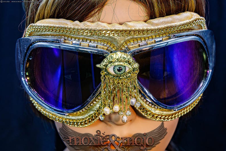 May include: Goggles with dark purple lenses and a silver frame, featuring a gold-toned trim with chain accents. A central gold ornament depicts an eye with dangling pearl and crystal charms. The text moxie & mojo is visible at the bottom.