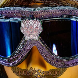 Puede incluir: Gafas protectoras moradas con un emblema de flor de loto en plata met&aacute;lica centrado en el puente. El marco est&aacute; adornado con peque&ntilde;os cristales oscuros y un borde texturizado. Las lentes son oscuras con un reflejo azul, texto moxie & mojo visible.