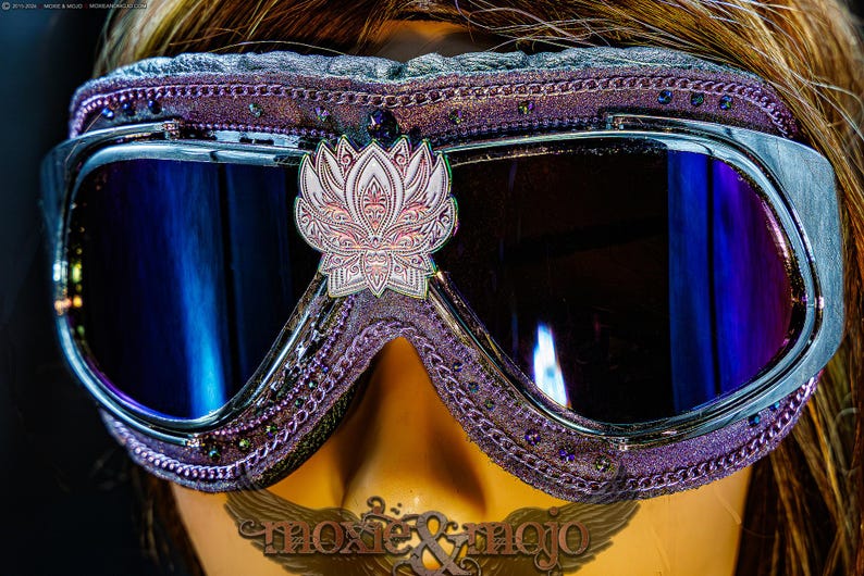 Puede incluir: Gafas protectoras moradas con un emblema de flor de loto en plata met&aacute;lica centrado en el puente. El marco est&aacute; adornado con peque&ntilde;os cristales oscuros y un borde texturizado. Las lentes son oscuras con un reflejo azul, texto moxie & mojo visible.