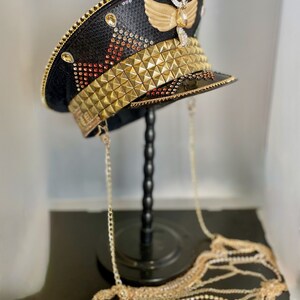 May include: A black and gold embellished hat with a gold chain. The hat has a gold winged design on the top and a gold studded band around the crown. The hat is on a black stand.
