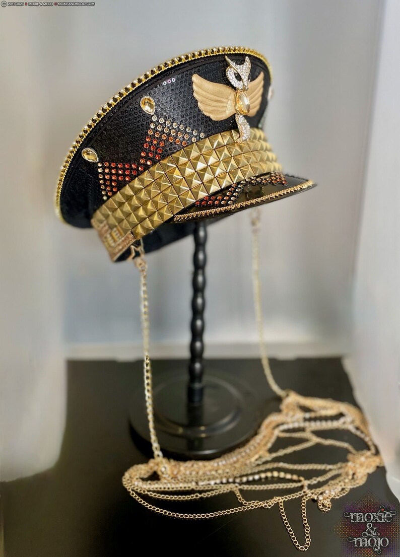May include: A black and gold embellished hat with a gold chain. The hat has a gold winged design on the top and a gold studded band around the crown. The hat is on a black stand.