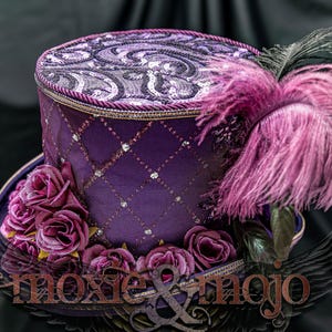 May include: A purple top hat featuring a diamond-patterned overlay with small silver accents, a top panel with a swirling sequin design, a cluster of dark purple fabric roses at the base, and a large pink feather plume. The text moxie & mojo is visible.