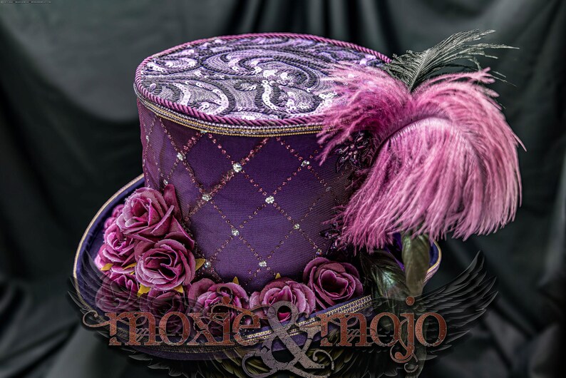 May include: A purple top hat featuring a diamond-patterned overlay with small silver accents, a top panel with a swirling sequin design, a cluster of dark purple fabric roses at the base, and a large pink feather plume. The text moxie & mojo is visible.