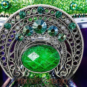 Pu&ograve; includere: Una spilla in metallo color argento con una grande pietra ovale verde dalla consistenza screpolata al centro. Il pezzo &egrave; decorato con strass verdi circolari pi&ugrave; piccoli disposti a ventaglio sopra la pietra centrale. Il testo moxie & mojo &egrave; visibile in basso.