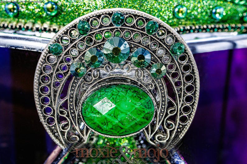 Pu&ograve; includere: Una spilla in metallo color argento con una grande pietra ovale verde dalla consistenza screpolata al centro. Il pezzo &egrave; decorato con strass verdi circolari pi&ugrave; piccoli disposti a ventaglio sopra la pietra centrale. Il testo moxie & mojo &egrave; visibile in basso.