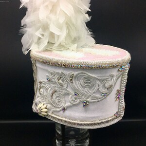 May include: A white drum major hat with a white feather plume. The hat is decorated with silver embroidery and rhinestones. The hat is on a black background.