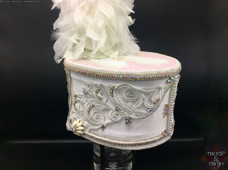 May include: A white drum major hat with a white feather plume. The hat is decorated with silver embroidery and rhinestones. The hat is on a black background.