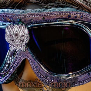 Puede incluir: Gafas protectoras de color morado con lentes oscuras y un emblema de flor de loto plateado centrado en el puente. El marco est&aacute; decorado con peque&ntilde;os cristales morados y adornos met&aacute;licos, con el texto moxie & mojo visible en la parte inferior.