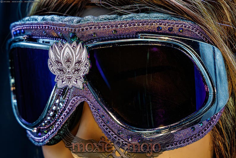 Puede incluir: Gafas protectoras de color morado con lentes oscuras y un emblema de flor de loto plateado centrado en el puente. El marco est&aacute; decorado con peque&ntilde;os cristales morados y adornos met&aacute;licos, con el texto moxie & mojo visible en la parte inferior.