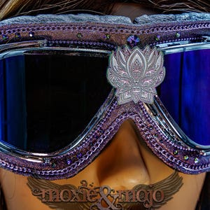 Puede incluir: Gafas protectoras de color morado con lentes oscuras reflectantes y marco met&aacute;lico plateado. El borde acolchado est&aacute; decorado con peque&ntilde;os brillantes morados y un emblema central de loto plateado. El texto moxie & mojo es visible.