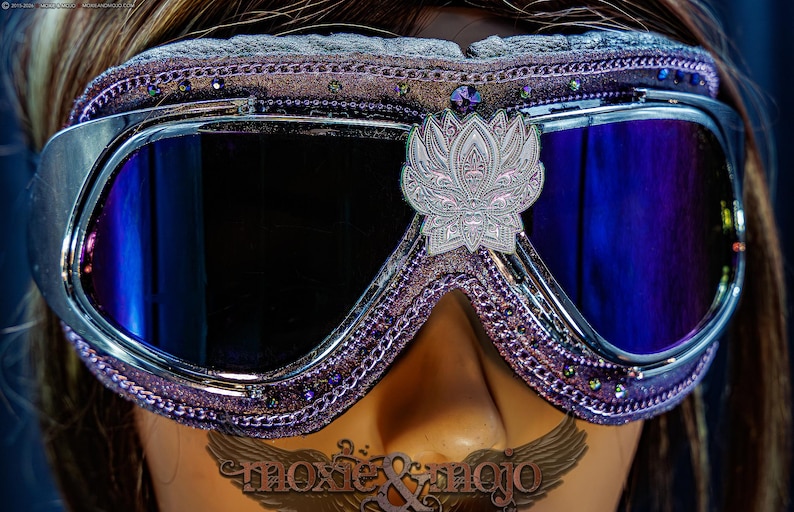 Puede incluir: Gafas protectoras de color morado con lentes oscuras reflectantes y marco met&aacute;lico plateado. El borde acolchado est&aacute; decorado con peque&ntilde;os brillantes morados y un emblema central de loto plateado. El texto moxie & mojo es visible.