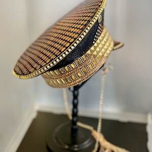 May include: A black and gold sequined military style hat with a gold chain attached. The hat is on a black stand.
