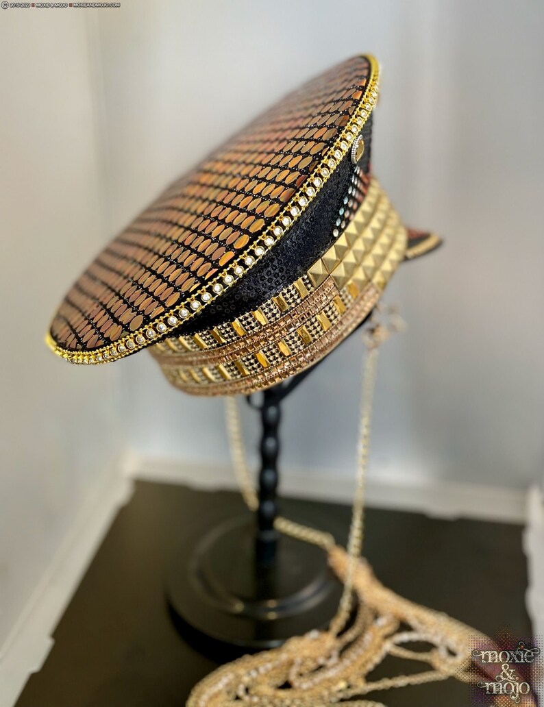 May include: A black and gold sequined military style hat with a gold chain attached. The hat is on a black stand.