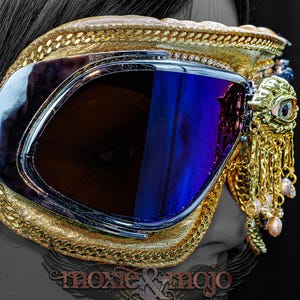 May include: Gold-trimmed goggles featuring dark blue reflective lenses and a brown elastic strap. The frame is decorated with a gold-toned eye emblem and dangling gold chains with small light-colored beads. The text moxie & mojo is visible.