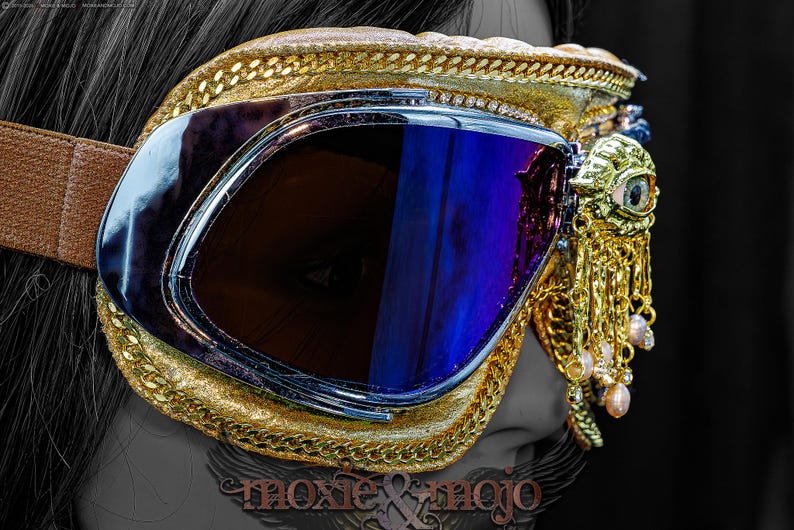 May include: Gold-trimmed goggles featuring dark blue reflective lenses and a brown elastic strap. The frame is decorated with a gold-toned eye emblem and dangling gold chains with small light-colored beads. The text moxie & mojo is visible.