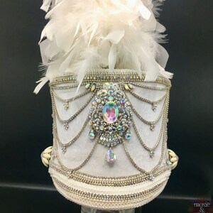 May include: White feather plume hat with a white velvet base and a large iridescent rhinestone embellishment. The hat is decorated with rows of silver chain and rhinestones.