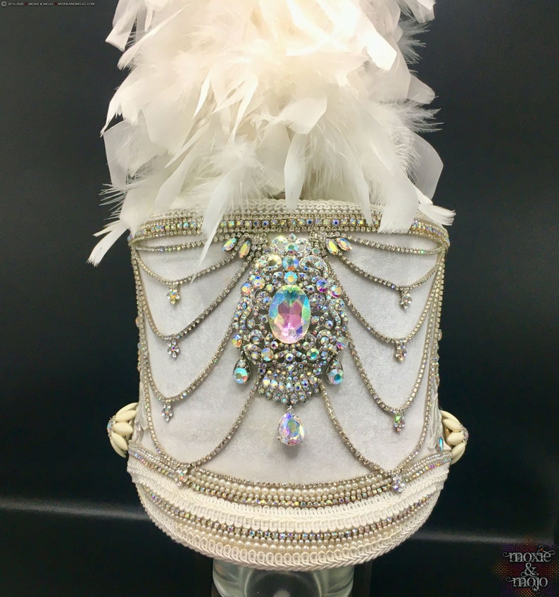 May include: White feather plume hat with a white velvet base and a large iridescent rhinestone embellishment. The hat is decorated with rows of silver chain and rhinestones.