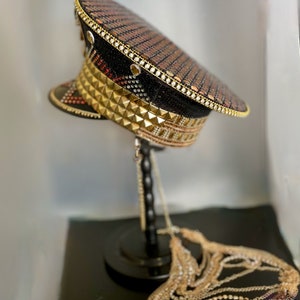 May include: A black and gold sequined hat with gold pyramid studs and a chain attached. The hat is on a black stand.