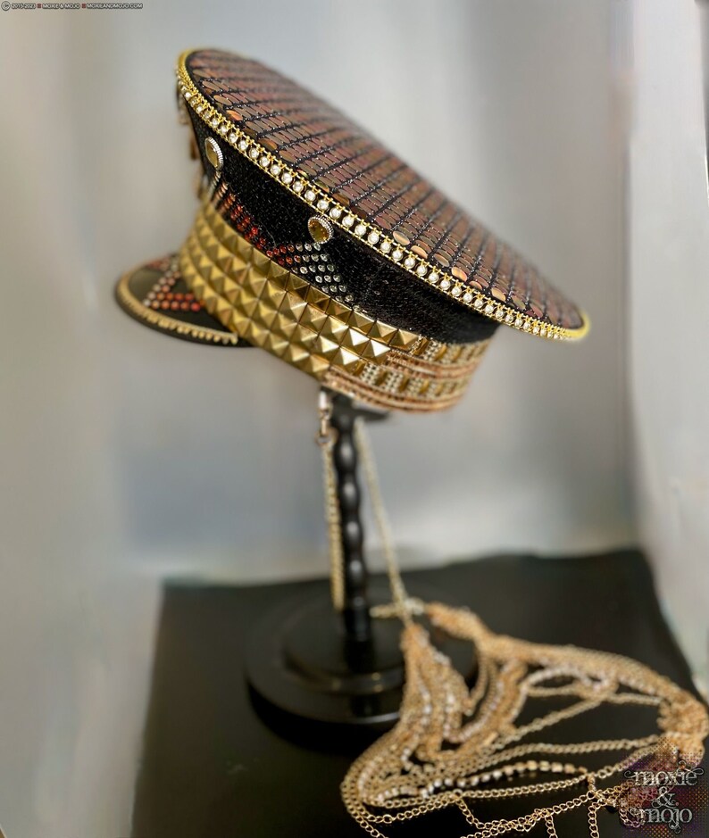 May include: A black and gold sequined hat with gold pyramid studs and a chain attached. The hat is on a black stand.