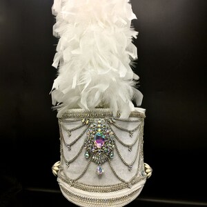 May include: A white feather plume atop a white hat with a silver and crystal embellishment. The hat is decorated with a large iridescent crystal and silver chains.