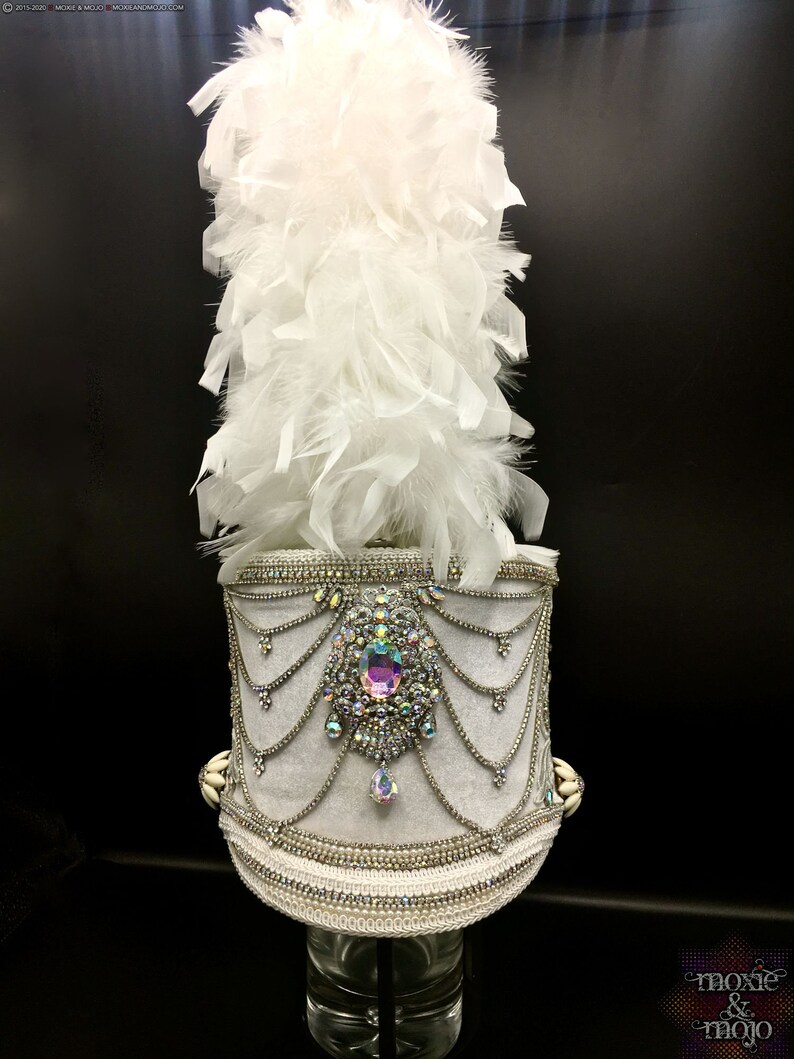 May include: A white feather plume atop a white hat with a silver and crystal embellishment. The hat is decorated with a large iridescent crystal and silver chains.