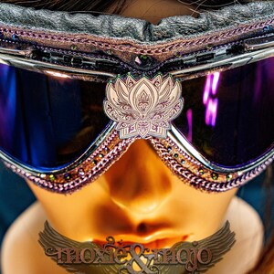 Puede incluir: Gafas protectoras con lentes reflectantes de color morado oscuro y un emblema central de flor de loto blanca. El marco presenta adornos en rosa met&aacute;lico, acolchado de cuero negro y peque&ntilde;as cuentas oscuras en los bordes. Texto moxie & mojo visible.