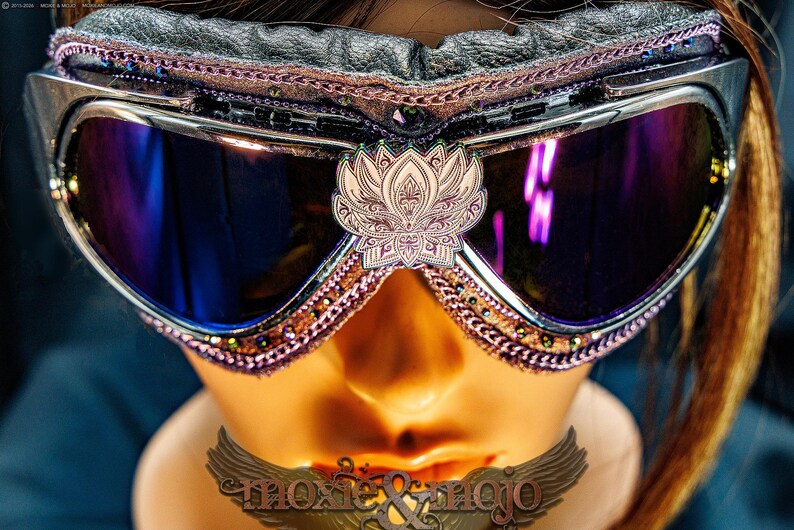 Puede incluir: Gafas protectoras con lentes reflectantes de color morado oscuro y un emblema central de flor de loto blanca. El marco presenta adornos en rosa met&aacute;lico, acolchado de cuero negro y peque&ntilde;as cuentas oscuras en los bordes. Texto moxie & mojo visible.