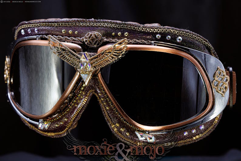 K&ouml;nnte beinhalten: Dunkelbraune Schutzbrille mit kupferfarbenem Metallrahmen und dunklen Gl&auml;sern. Der Steg ist mit einem goldenen Adleremblem mit Kristallakzenten verziert. Der Rahmen ist mit kleinen goldenen Nieten, klaren Kristallen und goldenen Ketten verziert.
