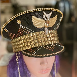May include: A black and gold sequined hat with a gold studded band and a winged angel design. The hat is decorated with rhinestones and has a gold chain attached to the side.
